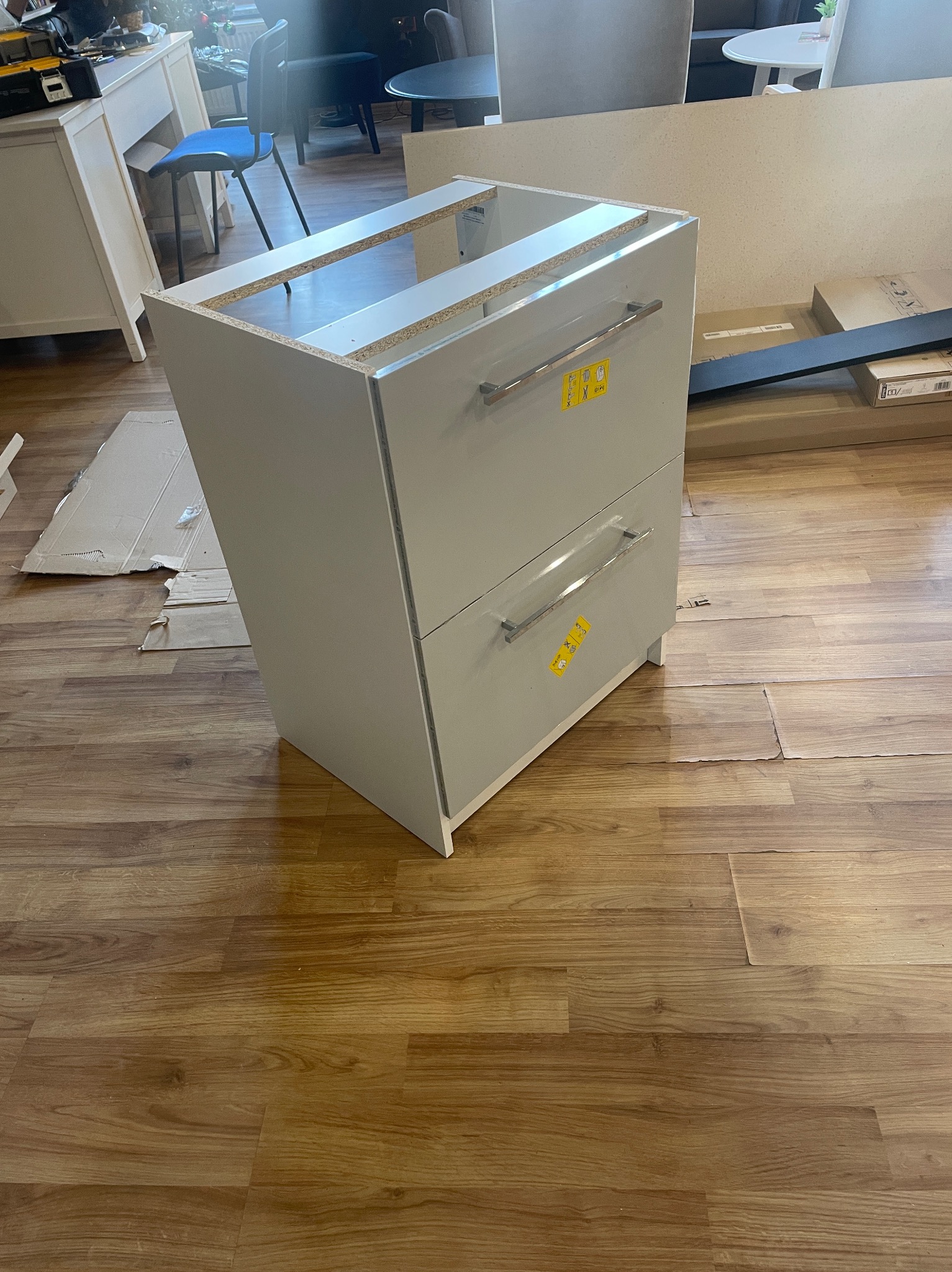 Niedokończona biała szafka z dwoma szufladami i srebrnymi uchwytami, z naklejkami ostrzegawczymi, stoi na podłodze z paneli imitujących drewno. Szafka bez blatu, w tle widoczne inne meble i kartony.