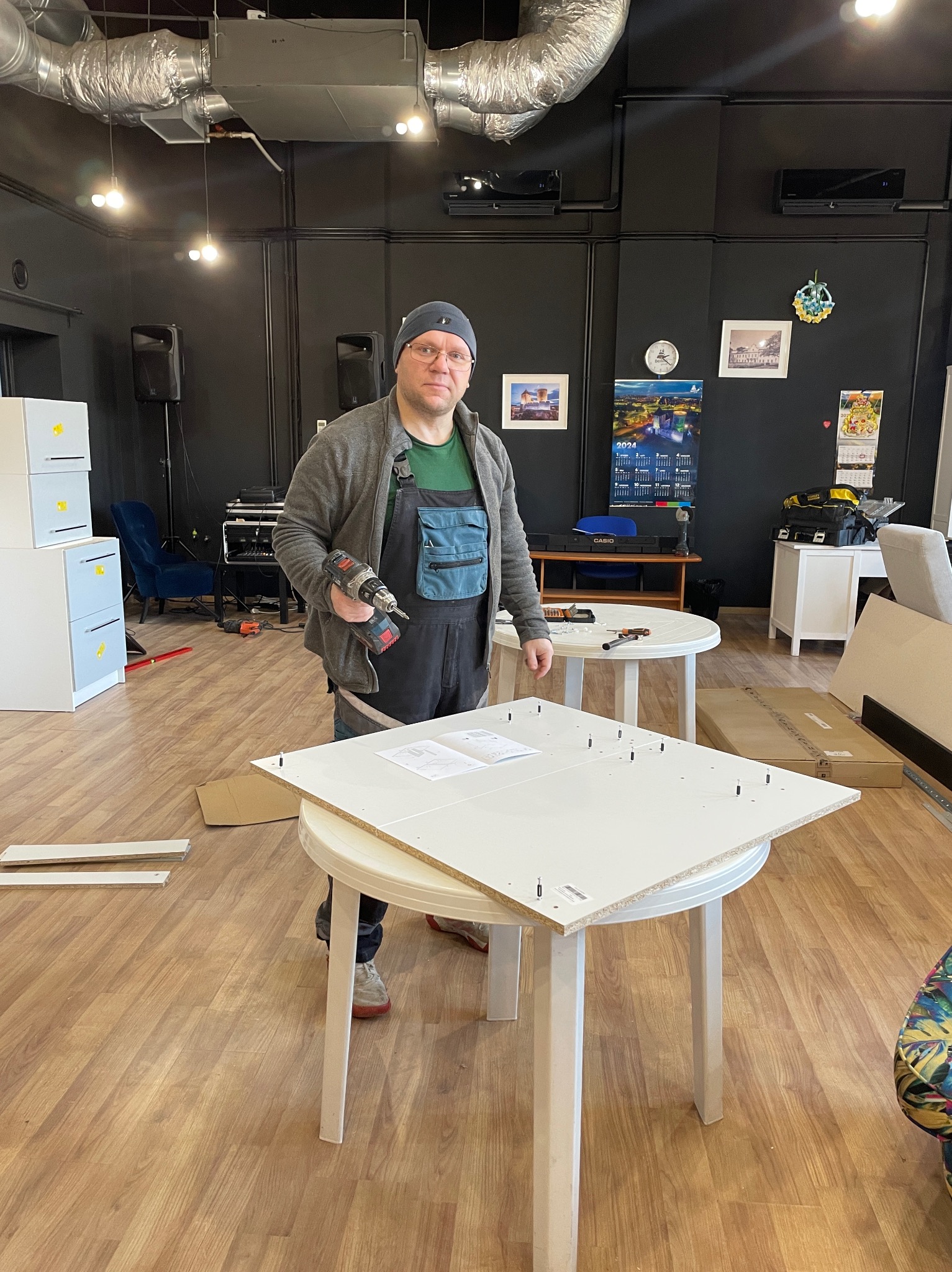 Mężczyzna w okularach i czapce, ubrany w roboczy kombinezon, trzyma wiertarkę akumulatorową nad białą płytą meblową z kołkami, leżącą na okrągłym stole. W tle widoczne są elementy wyposażenia...