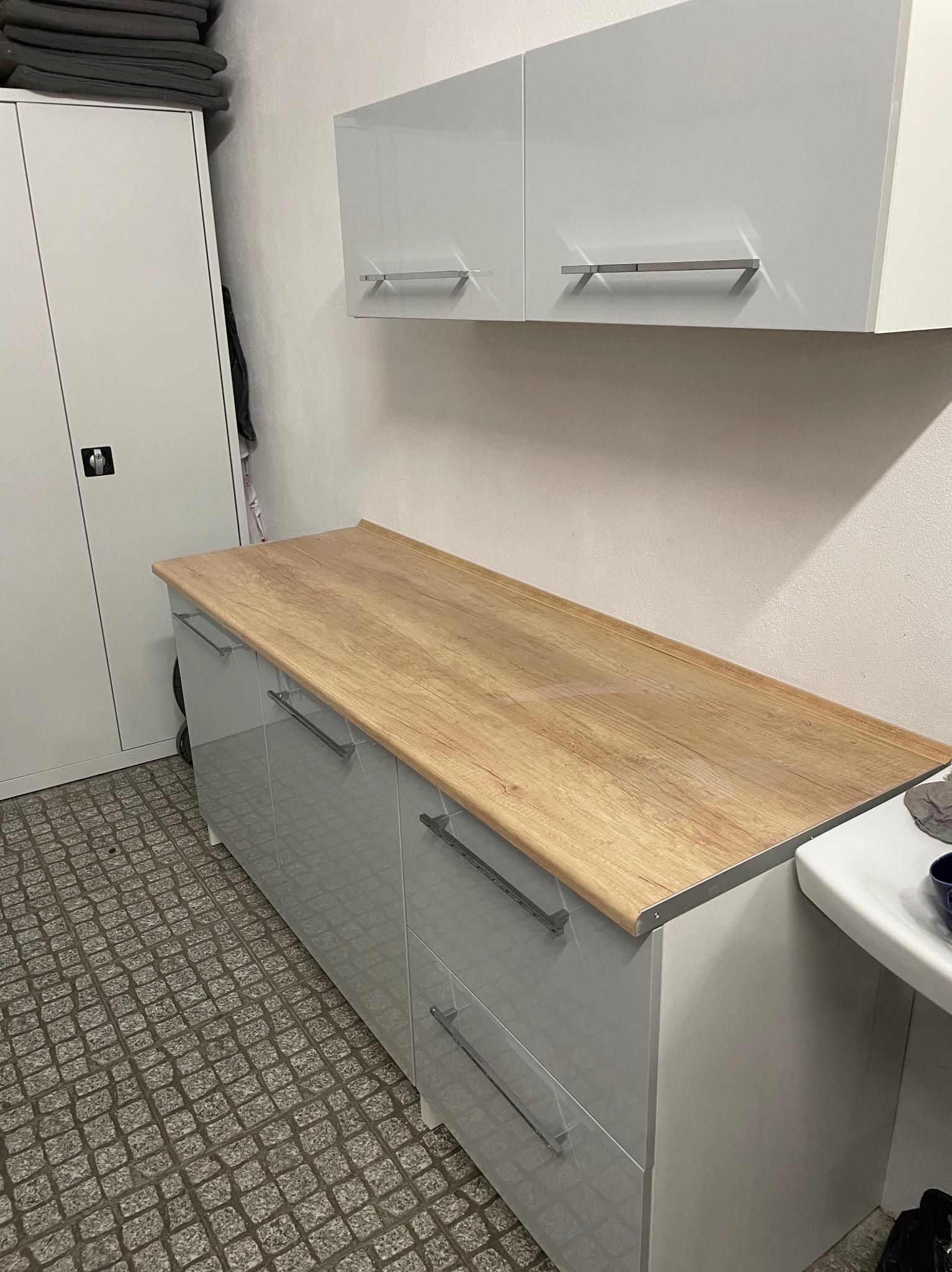 Szare szafki z drewnianym blatem i wiszące szafki w łazience, widoczne metalowe uchwyty i kostka brukowa na podłodze.