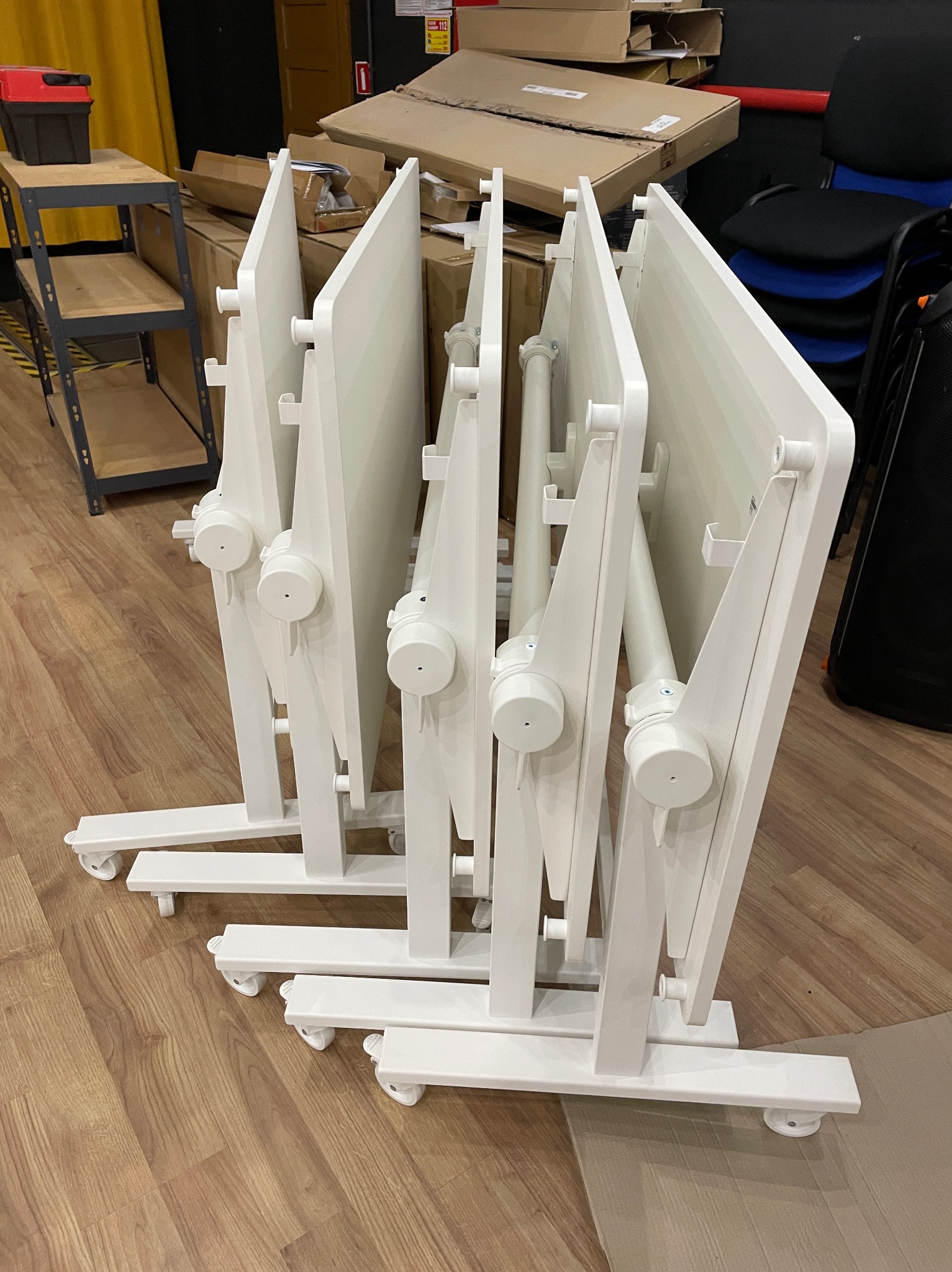 Składane, białe stoły konferencyjne na kółkach, ustawione jeden za drugim w magazynie, widoczne kartony i regał z narzędziami w tle.