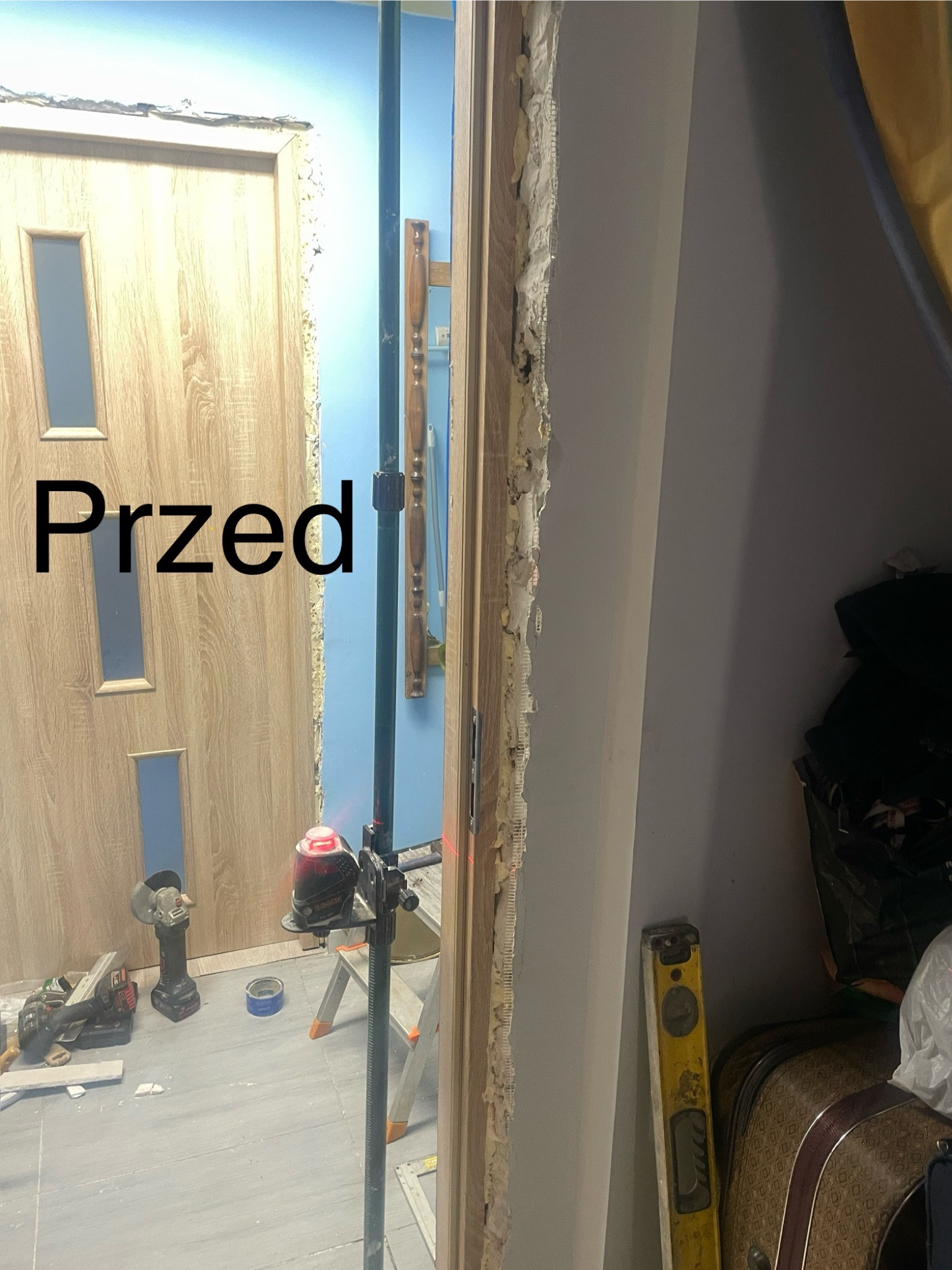 Demontaż ościeżnicy drzwiowej, widoczne narzędzia: szlifierka kątowa, poziomica, laser krzyżowy, fragment drzwi z okleiną drewnopodobną, ślady po starej ościeżnicy.