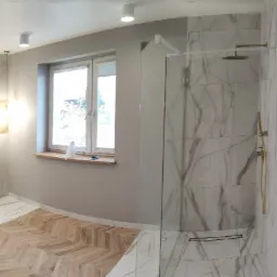 Nowoczesna łazienka z kabiną prysznicową typu walk-in, umywalką i bidetem, wykończona płytkami imitującymi marmur i drewnianym parkietem ułożonym we wzór jodełki, oświetlona punktowymi lampami...