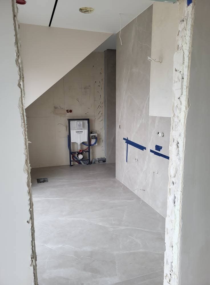 Wnętrze łazienki w trakcie wykończenia: szare płytki imitujące marmur na podłodze i ścianach, widoczna instalacja podtynkowa WC, surowy stan wykończenia przy wejściu.