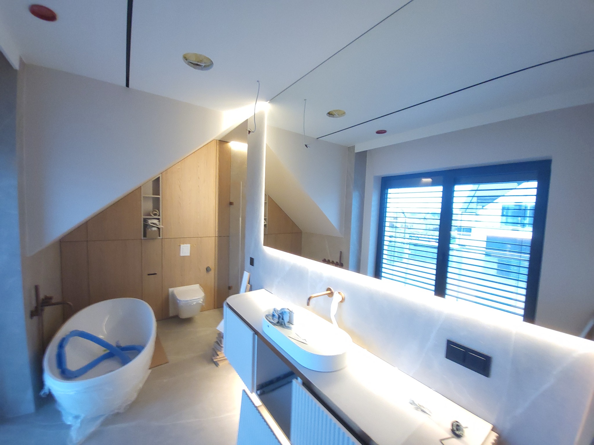 Nowoczesna łazienka w trakcie wykończenia: biała wanna w folii, umywalka z podświetleniem LED, drewniane szafki i okno z żaluzjami. Widoczne elementy instalacji.