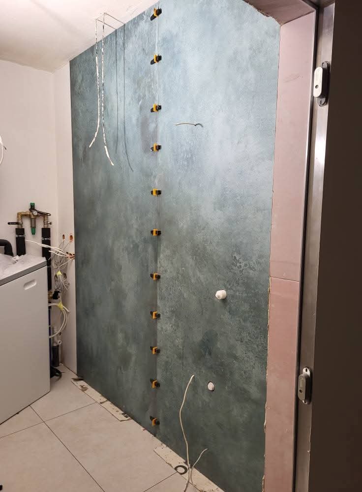 Ściana w łazience z efektem dekoracyjnym, zabezpieczona pionowymi listwami z klipsami. Widoczne instalacje i fragmenty urządzeń sanitarnych.