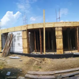 Budowa domu jednorodzinnego: ściany z bloczków betonowych, drewniane szalunki stropu, zbrojenie wystające ponad ściany, widoczna izolacja fundamentów od strony zewnętrznej.