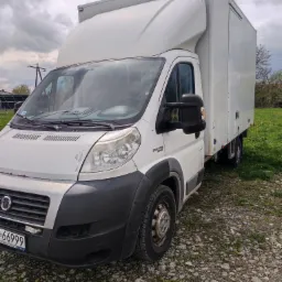 Biały samochód dostawczy Fiat Ducato z zabudową kontenerową, zaparkowany na żwirowym podłożu z widocznymi śladami użytkowania na karoserii.