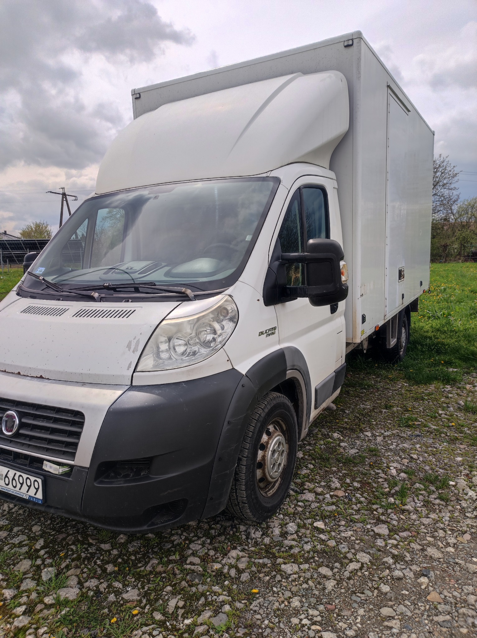 Biały samochód dostawczy Fiat Ducato z zabudową kontenerową, zaparkowany na żwirowym podłożu z widocznymi śladami użytkowania na karoserii.