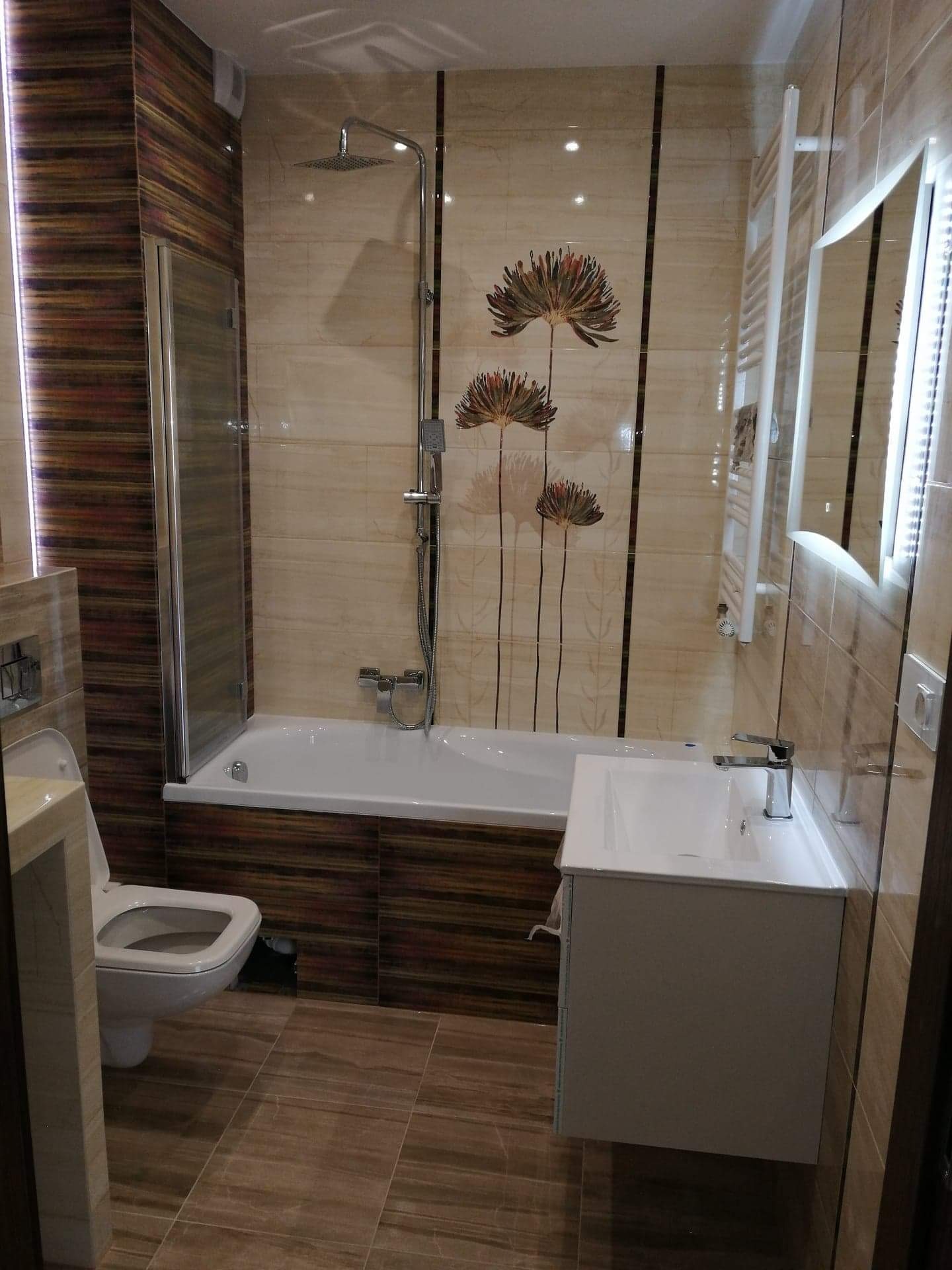 Wykończona łazienka z beżowo-brązowymi płytkami imitującymi drewno, z kabiną prysznicową nad wanną, umywalką nablatową i dekoracyjnym motywem kwiatowym na ścianie.
