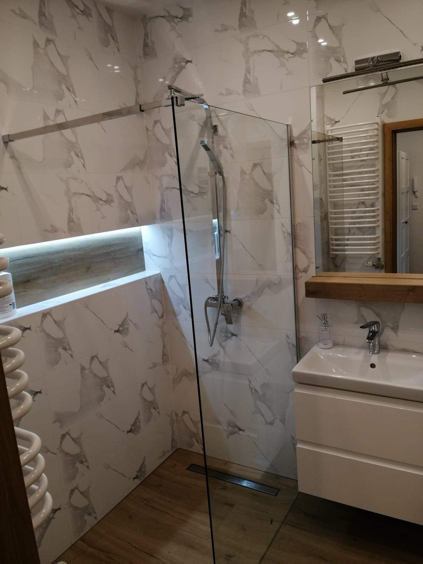 Nowoczesna łazienka z kabiną prysznicową typu walk-in, wykończona płytkami imitującymi marmur, drewnopodobną podłogą, z podświetlaną wnęką i minimalistyczną umywalką nablatową.