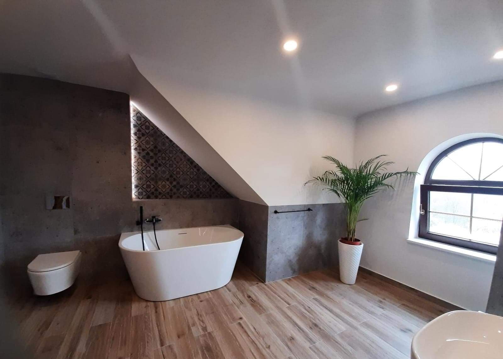 Nowoczesna łazienka na poddaszu z wolnostojącą wanną, drewnopodobną podłogą, szarą ścianą z dekoracyjnym akcentem mozaiki, rośliną doniczkową w białej osłonce i oknem łukowym.