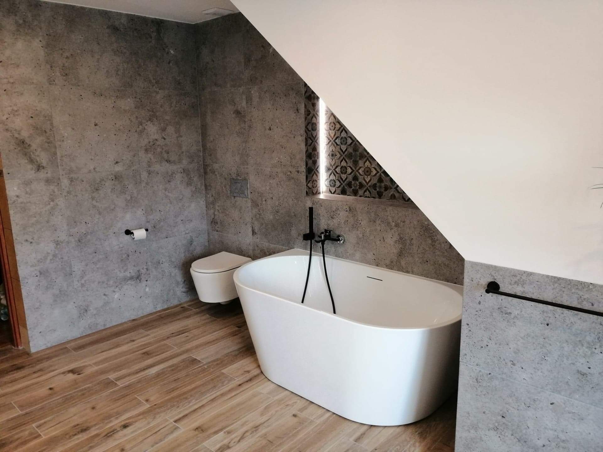 Nowoczesna łazienka na poddaszu z wolnostojącą białą wanną, toaletą podwieszaną i szarą okładziną ścian imitującą beton. Uwagę zwraca skośny sufit i fragment dekoracyjnej mozaiki w trójkątnym oknie.