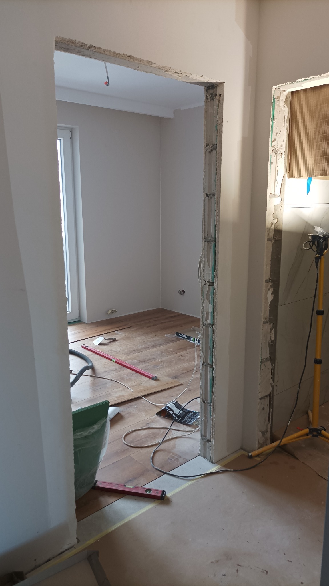 Demontaż ościeżnicy drzwiowej w trakcie remontu mieszkania, widoczne odsłonięte cegły, fragmenty płytek i drewniana podłoga z poziomnicą.