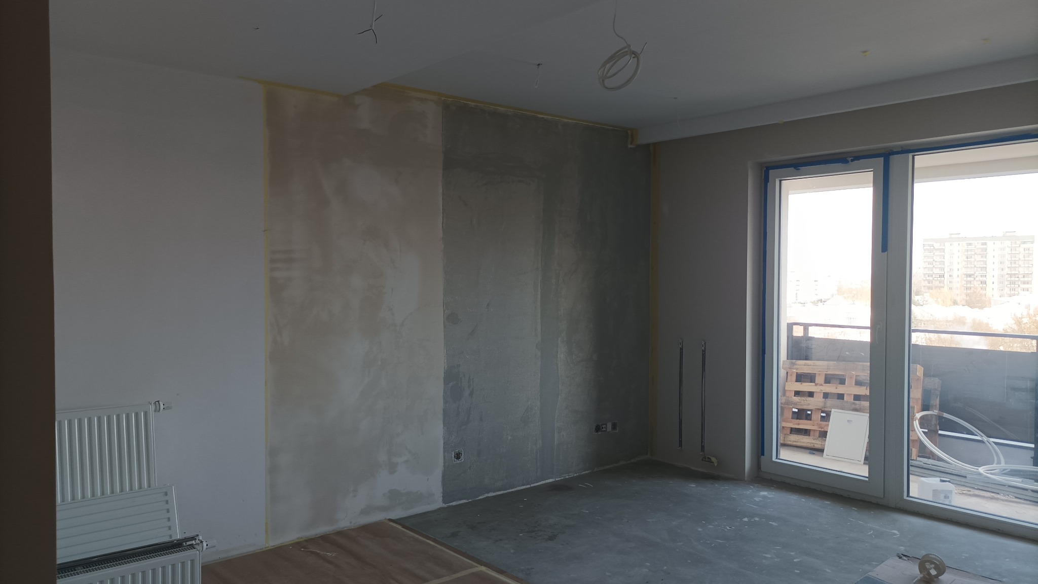 Wnętrze pokoju w trakcie prac wykończeniowych: ściany częściowo przygotowane pod malowanie, widoczne ślady szpachlowania i taśmy malarskie, grzejnik oraz okno balkonowe z widokiem na miasto.