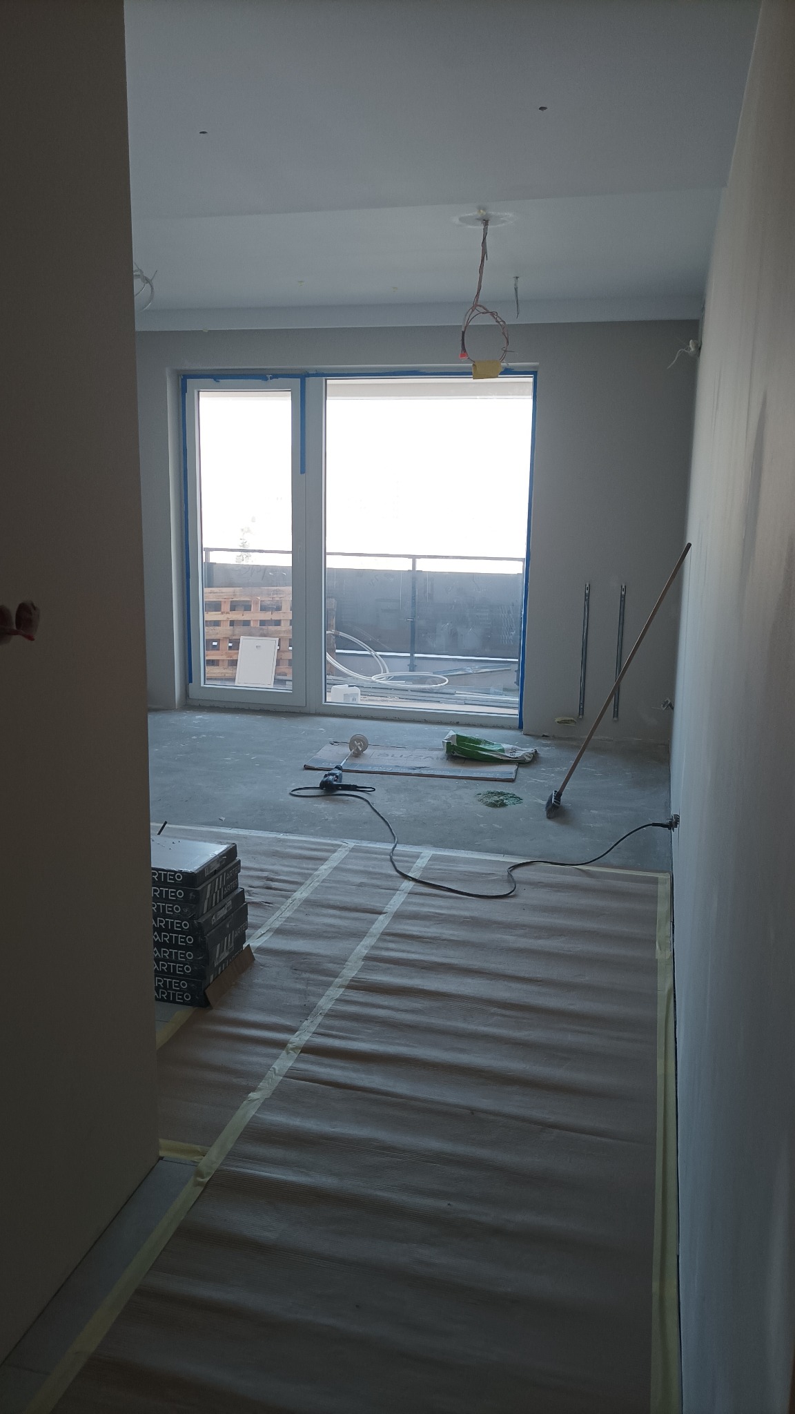 Wnętrze w trakcie prac przygotowawczych: podłoga zabezpieczona folią, ściany przygotowane do malowania, widoczne narzędzia i materiały budowlane, okno balkonowe z widokiem na zewnątrz.