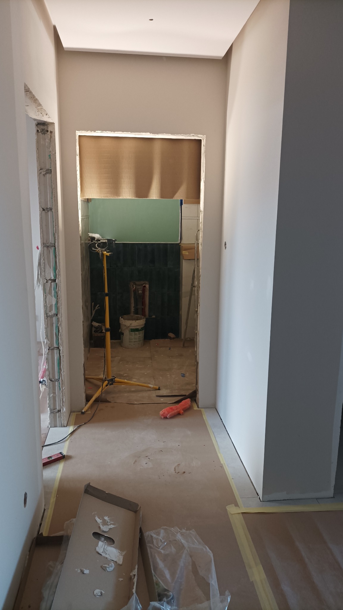 Korytarz w trakcie remontu, widoczne przygotowane ściany, podłoga zabezpieczona kartonem, w głębi pomieszczenie z ciemnozielonymi płytkami i lampą budowlaną.