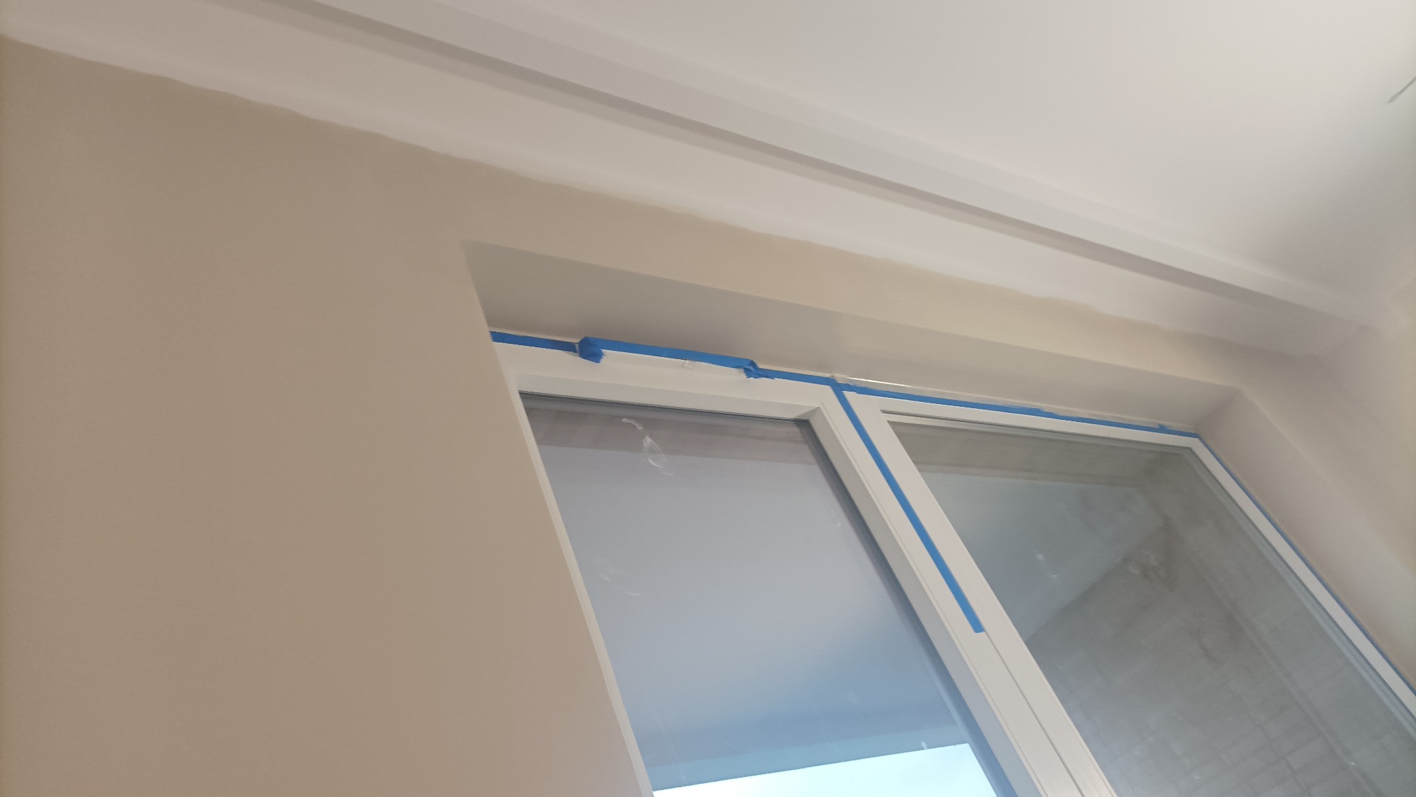 Ściana w trakcie malowania z taśmą malarską zabezpieczającą ramę okienną i listwę sufitową, widoczne niedociągnięcia w nałożonej warstwie farby.