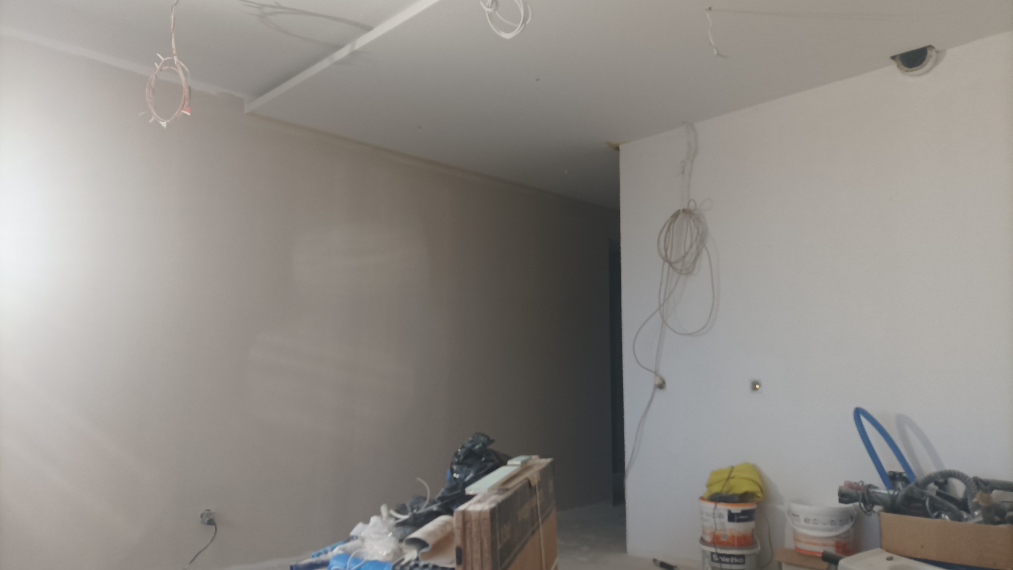 Wnętrze w trakcie prac wykończeniowych: widoczne gładkie, pomalowane ściany, częściowo zamontowany sufit podwieszany, wystające kable, narzędzia i materiały budowlane w nieładzie przy ścianie.
