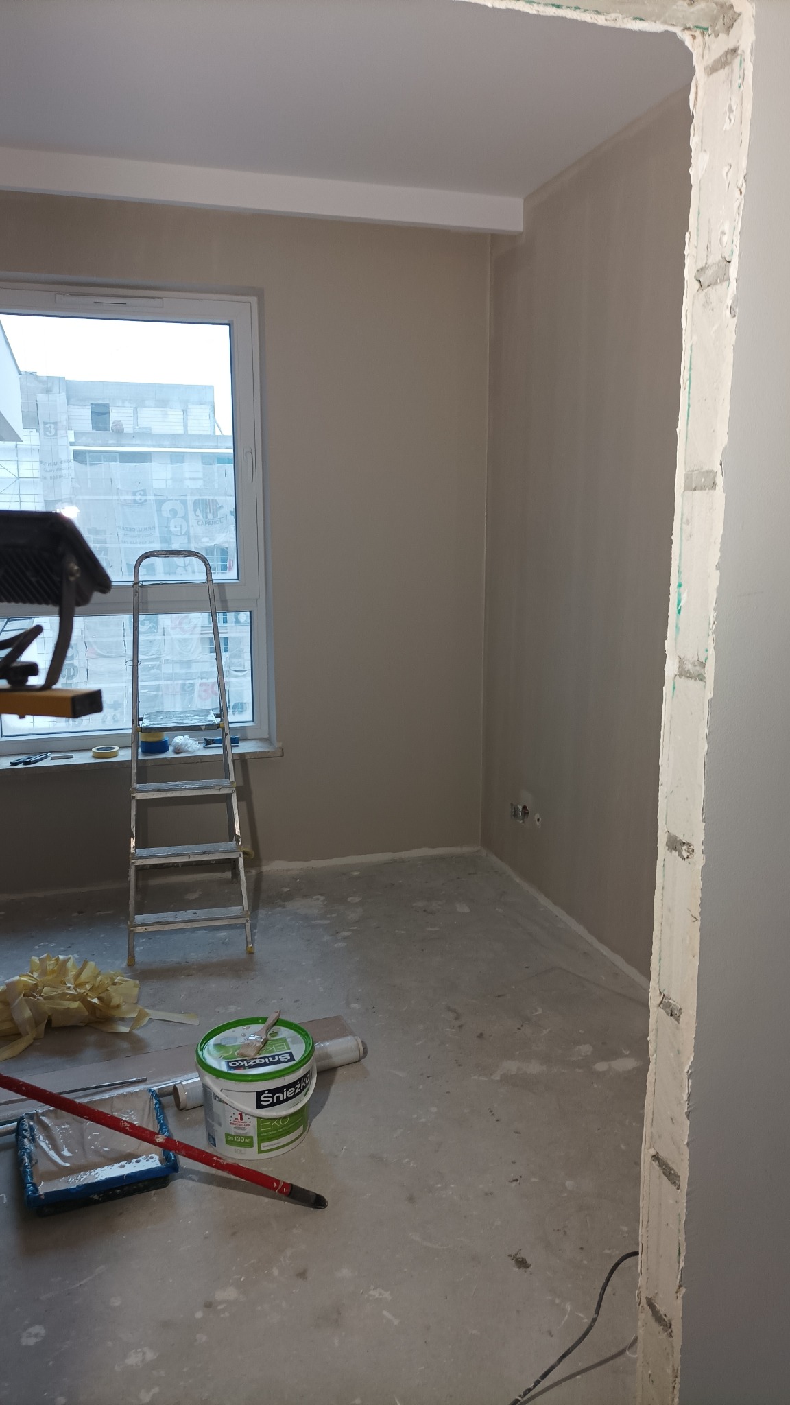 Pokój w trakcie malowania ścian na beżowo, widoczna drabina, wiadro z farbą Śnieżka EKO, wałek malarski, folia malarska, oraz fragment surowej ściany przy przejściu do innego pomieszczenia.