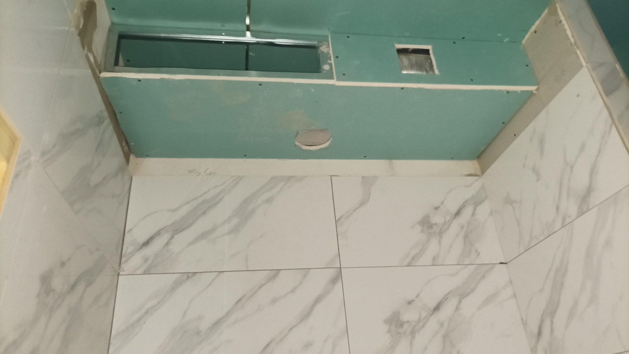 Wnętrze łazienki w trakcie prac wykończeniowych: położone białe płytki imitujące marmur na ścianach, sufit podwieszany z zielonych płyt kartonowo-gipsowych z otworem na oświetlenie i wentylację.