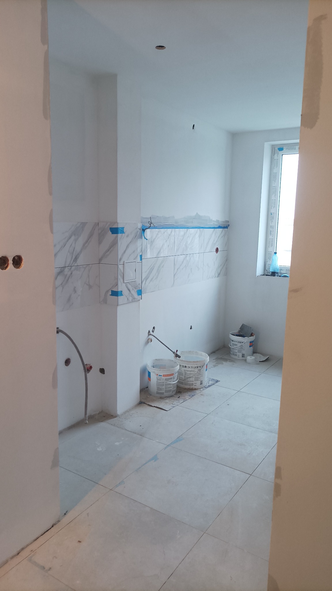 Łazienka w trakcie wykańczania: częściowo położone marmurowe płytki z niebieską taśmą zabezpieczającą, dwa wiadra z zaprawą na podłodze z jasnych płytek, widoczne przyłącza hydrauliczne.