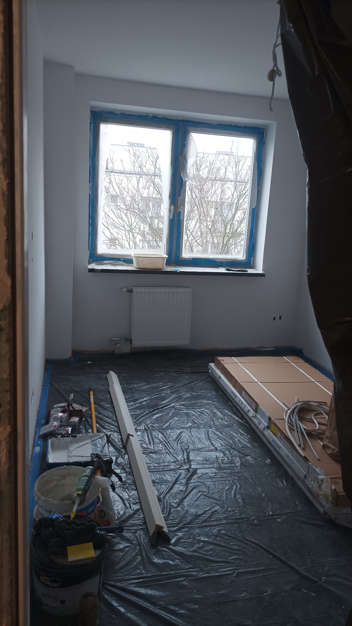 Pokój w trakcie remontu: zabezpieczona folią podłoga, okna z niebieską taśmą malarską, wiadro z farbą Dulux White, pędzle, listwy przypodłogowe i paczki paneli czekające na montaż.