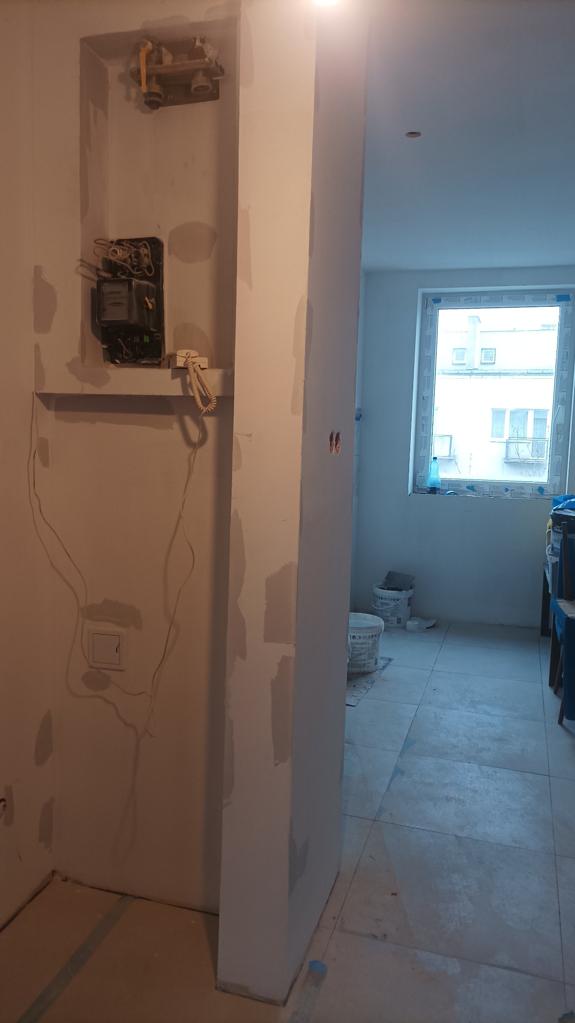 Wnętrze w trakcie remontu: ściany częściowo pokryte szpachlą, widoczny licznik elektryczny, puszka instalacyjna, wiadro z farbą w tle oraz fragment okna.