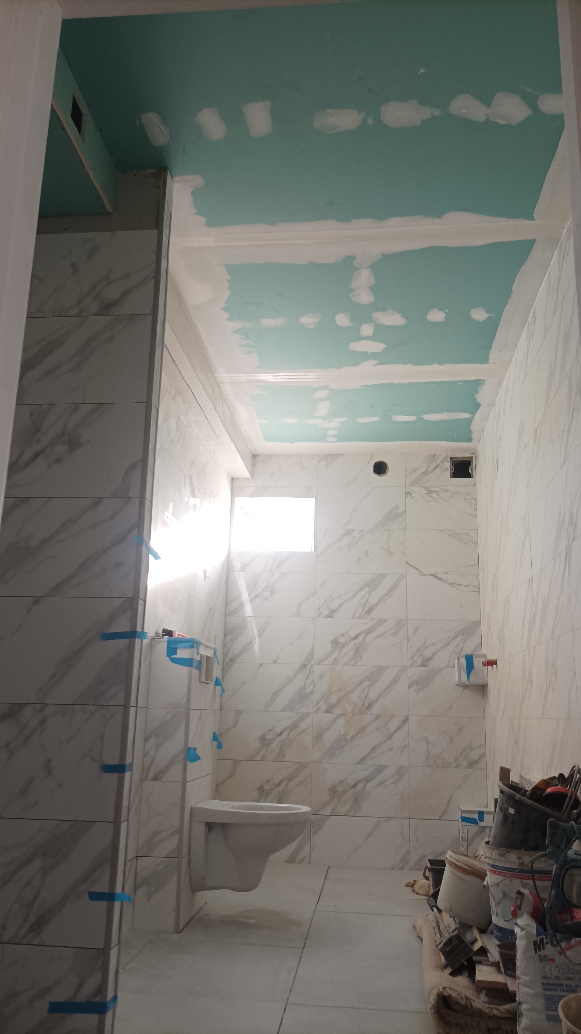 Łazienka w trakcie remontu: białe marmurowe płytki na ścianach, sufit pomalowany na turkusowo z widocznymi łatami szpachli, wisząca muszla klozetowa, narzędzia i materiały budowlane na podłodze.