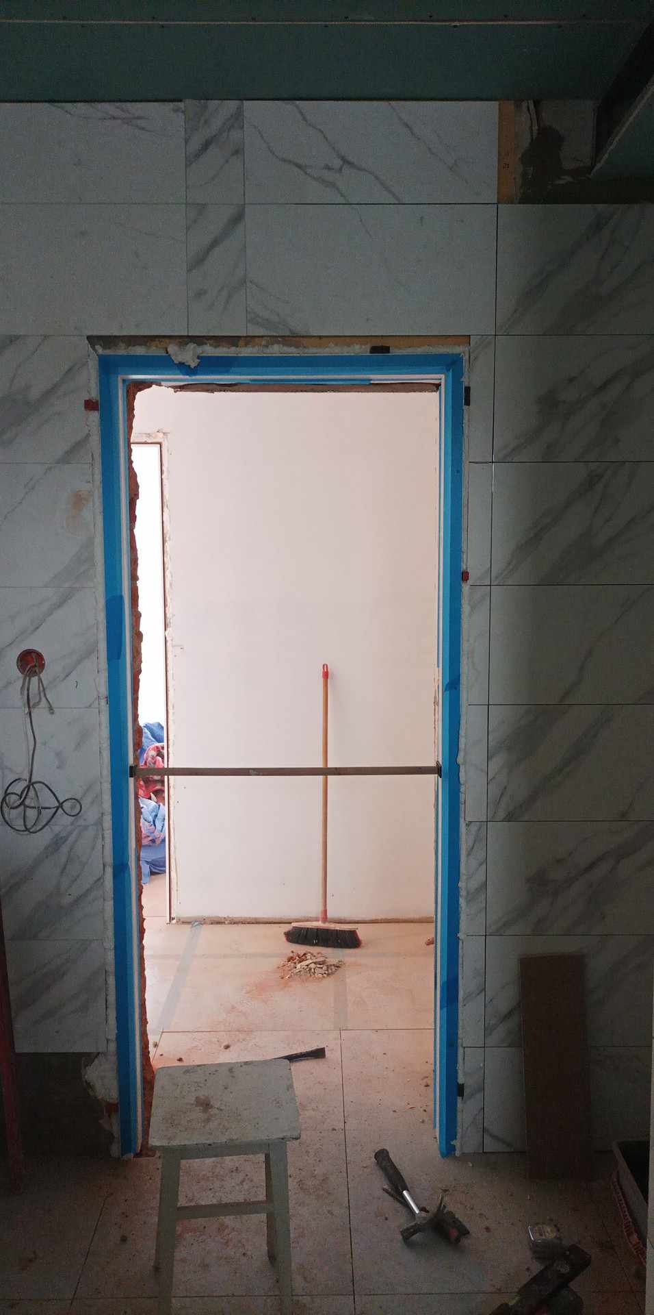Wnętrze w trakcie remontu: Ściany wyłożone marmurkowymi płytkami, zabezpieczone niebieską taśmą ościeżnice, widoczna biała ściana w sąsiednim pomieszczeniu, na podłodze leży miotła i resztki gruzu,...