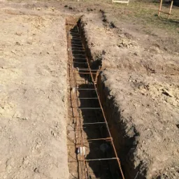 Wąski wykop w ziemi z ułożonym zbrojeniem stalowym, przygotowany pod wylanie fundamentu. W tle widoczne drewniane szalunki i stos bloczków betonowych.