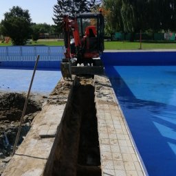 ADA MAX - Pomarańczowa mini koparka wykonuje wąski wykop wzdłuż krawędzi basenu z niebieską wodą, widoczny fragment drewnianego pala, w tle zieleń i ogrodzenie.