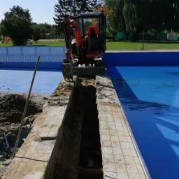 Pomarańczowa mini koparka wykonuje wąski wykop wzdłuż krawędzi basenu z niebieską wodą, widoczny fragment drewnianego pala, w tle zieleń i ogrodzenie.