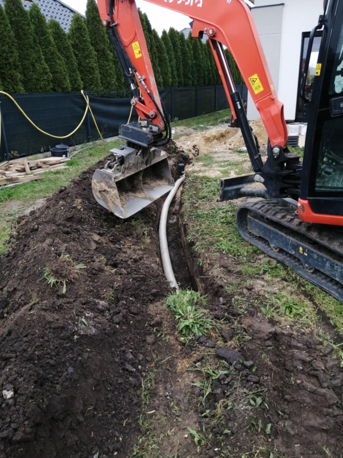 Pomarańczowa minikoparka Kubota z łyżką nad wykopem, w którym ułożono perforowaną rurę drenażową, w tle rząd tuj i ogrodzenie z siatki.