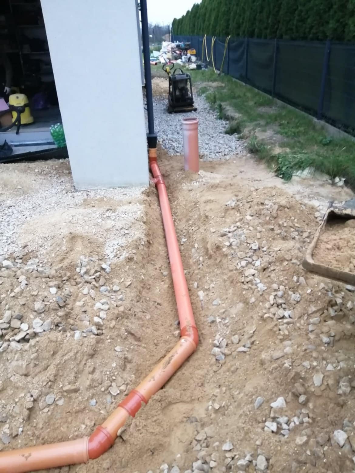 Pomarańczowa rura kanalizacyjna ułożona w wykopie z piasku i kamieni przy fundamencie budynku. W tle ubijarka wibracyjna i ogrodzenie z tujami.