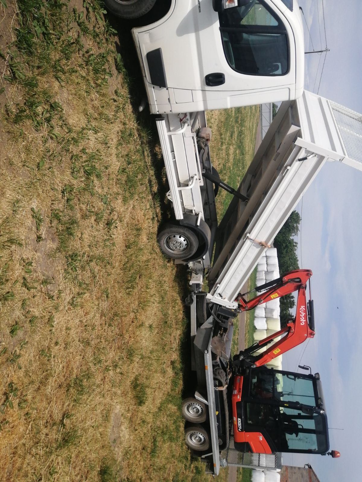 Biały samochód dostawczy z otwartą skrzynią ładunkową, na której transportowany jest czerwony minikoparka Kubota KX027-4, w tle widać bele słomy