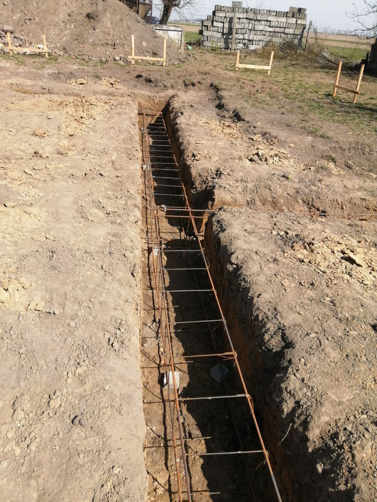 Wąski wykop w ziemi z ułożonym zbrojeniem stalowym, przygotowany pod wylanie fundamentu. W tle widoczne drewniane szalunki i stos bloczków betonowych.