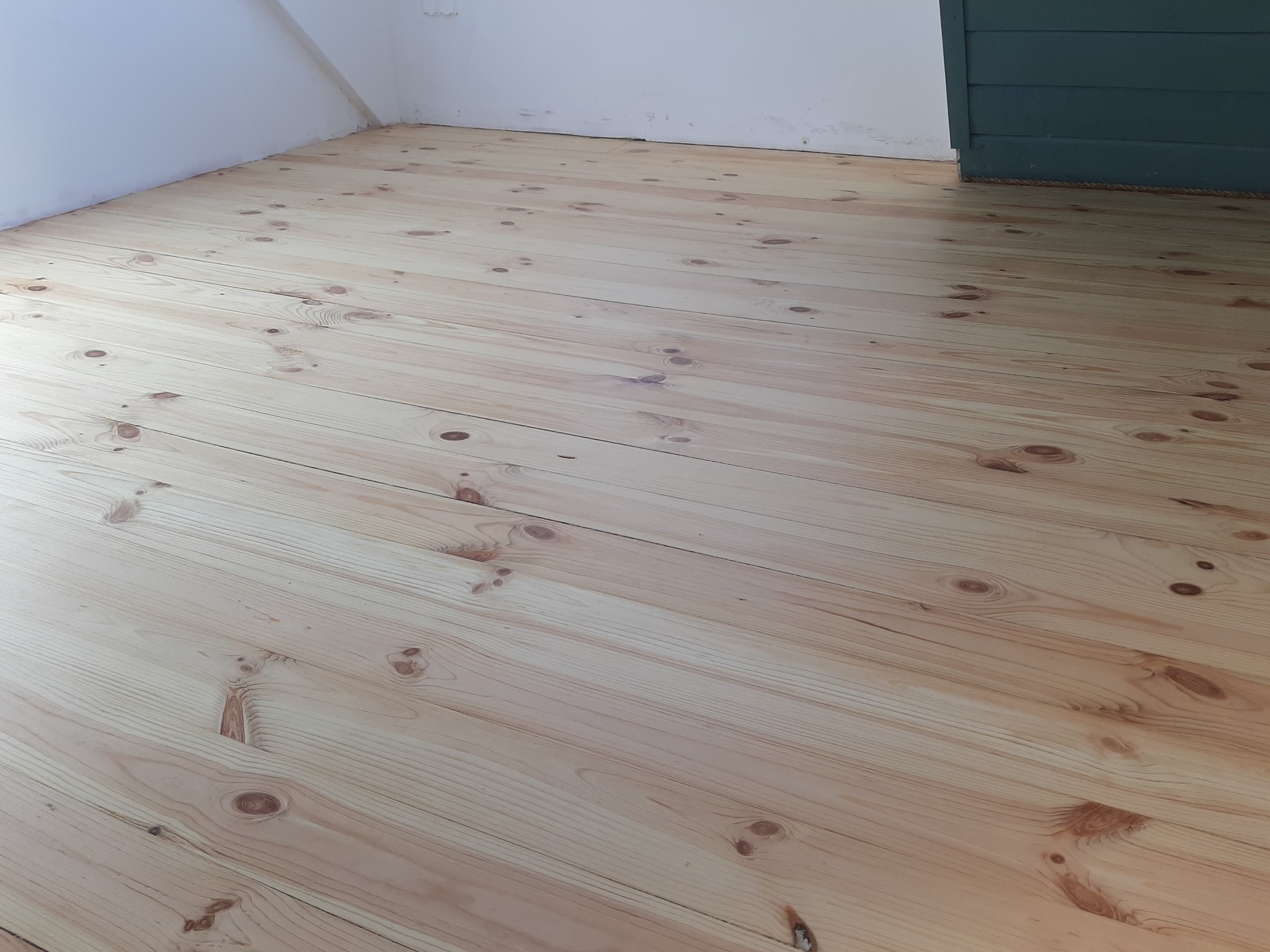 Jasna, świeżo położona podłoga z desek sosnowych z widocznymi sękami, ułożona wzdłuż pomieszczenia, z fragmentem zielonej boazerii w tle.