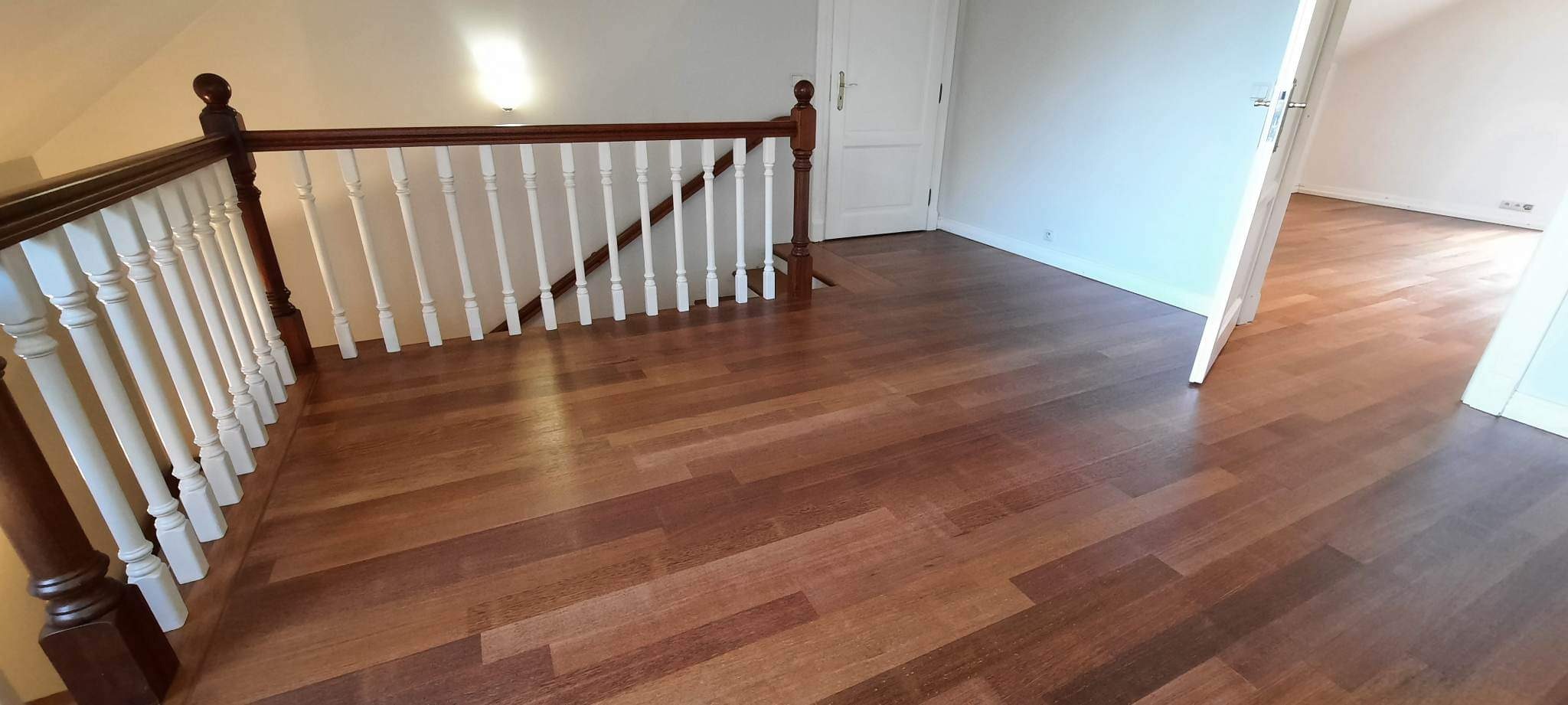 Wykończona podłoga z egzotycznego drewna ułożona w jodełkę francuską, widok z góry na balustradę schodów z białymi tralkami i ciemnobrązowymi słupkami, białe ściany i drzwi w tle.