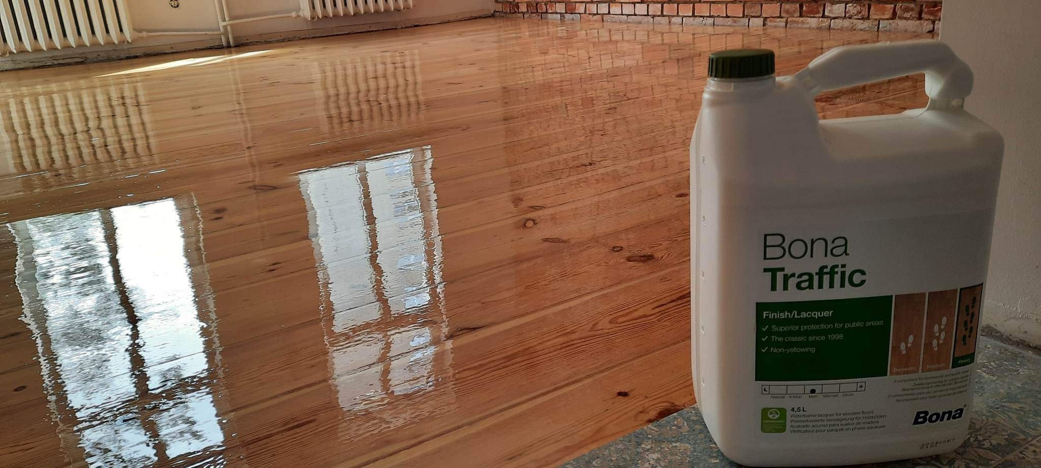 Świeżo polakierowana, błyszcząca drewniana podłoga z widocznym odbiciem okna i grzejnika; obok stoi kanister z lakierem Bona Traffic.