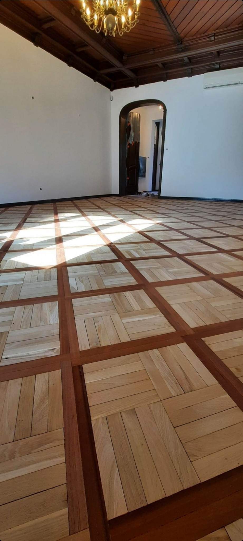 Klasyczny parkiet z jasnego drewna ułożony w kwadraty oddzielone ciemniejszymi listwami, widok na przestronne wnętrze z drewnianym sufitem i żyrandolem.