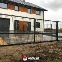 Nowoczesne ogrodzenie panelowe w kolorze grafitowym, z bramą wjazdową, na tle domu z elementami drewnianej elewacji i kostki brukowej, widok z poziomu gruntu.