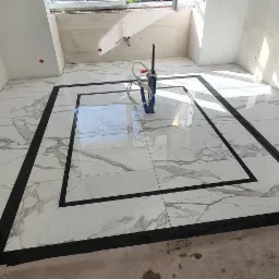 Układanie białych, marmurkowych płytek z czarną obwódką w pokoju z oknem, widoczne ślady po kleju na podłodze.