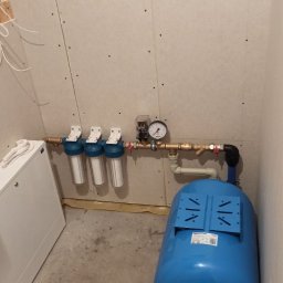 UTS Usługi Tymoteusz Soszka - Instalacja hydroforowa z trzema filtrami wody, manometrem i zbiornikiem ciśnieniowym w pomieszczeniu wykończonym płytami gipsowo-kartonowymi, widoczne elementy rur miedzianych i zawory.