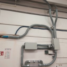 UTS Usługi Tymoteusz Soszka - Szare rury wentylacyjne z elementami przezroczystymi, zamontowane na ścianie w hali przemysłowej, z widocznymi puszkami przyłączeniowymi i fragmentami instalacji elektrycznej.