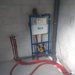 UTS Usługi Tymoteusz Soszka - Stelaż podtynkowy WC marki Roca zamontowany na tle surowych ścian z betonu komórkowego, widoczne czerwone rury instalacji wodnej i szara rura kanalizacyjna, całość zabezpieczona pianką montażową.