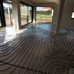 Rozłożona instalacja ogrzewania podłogowego na siatce w surowym wnętrzu domu z dużymi oknami tarasowymi i widokiem na ogród.