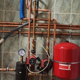 UTS Usługi Tymoteusz Soszka - Nowa instalacja centralnego ogrzewania z miedzianych rur, pomp obiegowych, czarnego filtra i czerwonego zbiornika wyrównawczego zamontowana na ścianie z szarych płytek.