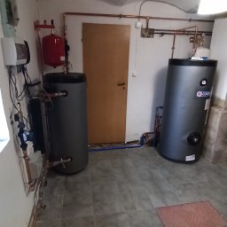 UTS Usługi Tymoteusz Soszka - Dwa szare zasobniki ciepłej wody użytkowej w kotłowni, z widocznymi rurami miedzianymi i elementami instalacji. Na podłodze szare płytki imitujące kamień.