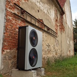 UTS Usługi Tymoteusz Soszka - Pompa ciepła powietrze-woda zamontowana na betonowym fundamencie przy starej, ceglanej ścianie budynku z widoczną drabiną.
