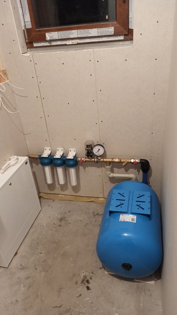 Instalacja hydroforowa z trzema filtrami wody, manometrem i zbiornikiem ciśnieniowym w pomieszczeniu wykończonym płytami gipsowo-kartonowymi, widoczne elementy rur miedzianych i zawory.